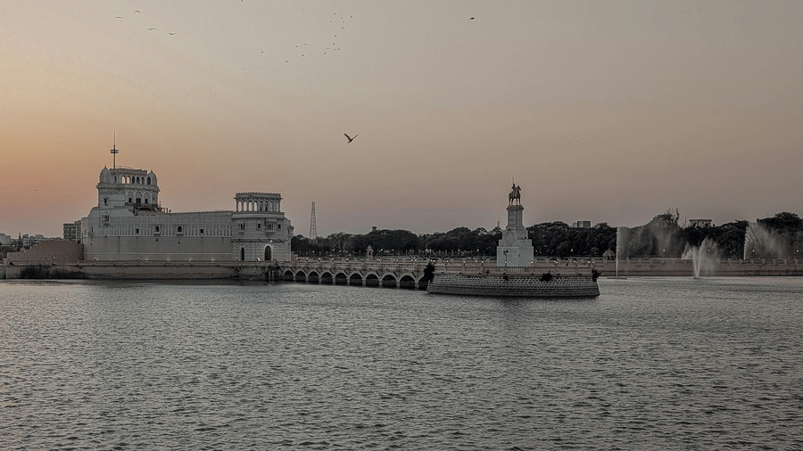 A view of a white building connected by a walkway over a lake, with another small white structure and surrounding buildings visible across the water.