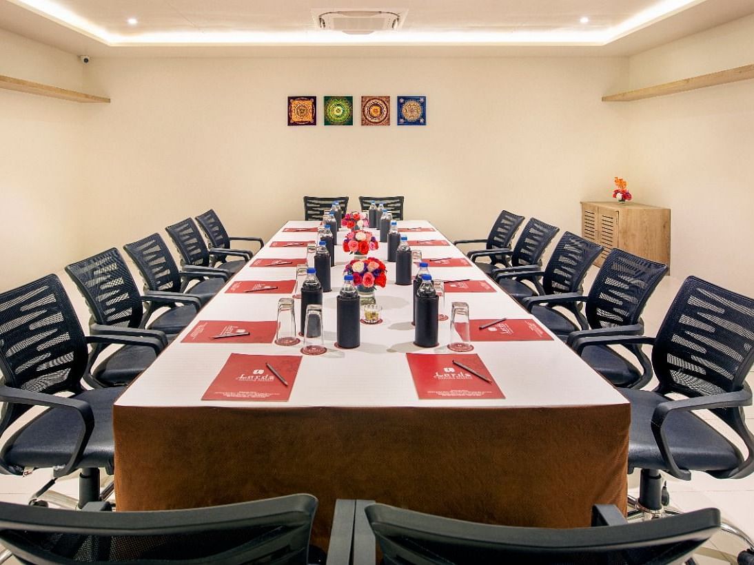 A formal meeting room is set up with a long rectangular table surrounded by numerous black office chairs, featuring water bottles and red and white placemats, under bright ceiling lights and with framed artwork on the wall - Lords Inn Motikhavdi, Jamnagar.