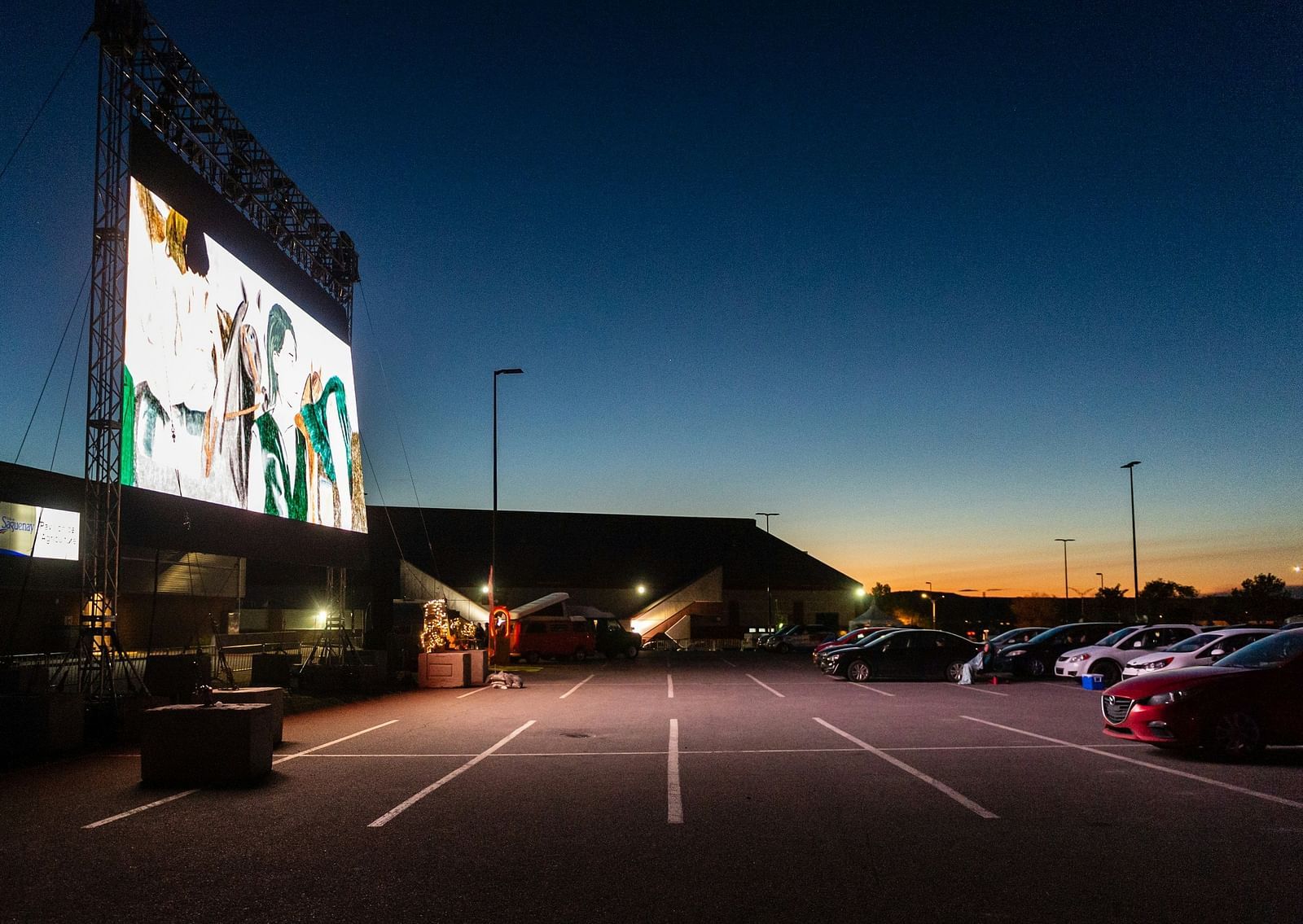 An overview of an outdoor cinema on a parking lot with a large screen and cars in view.