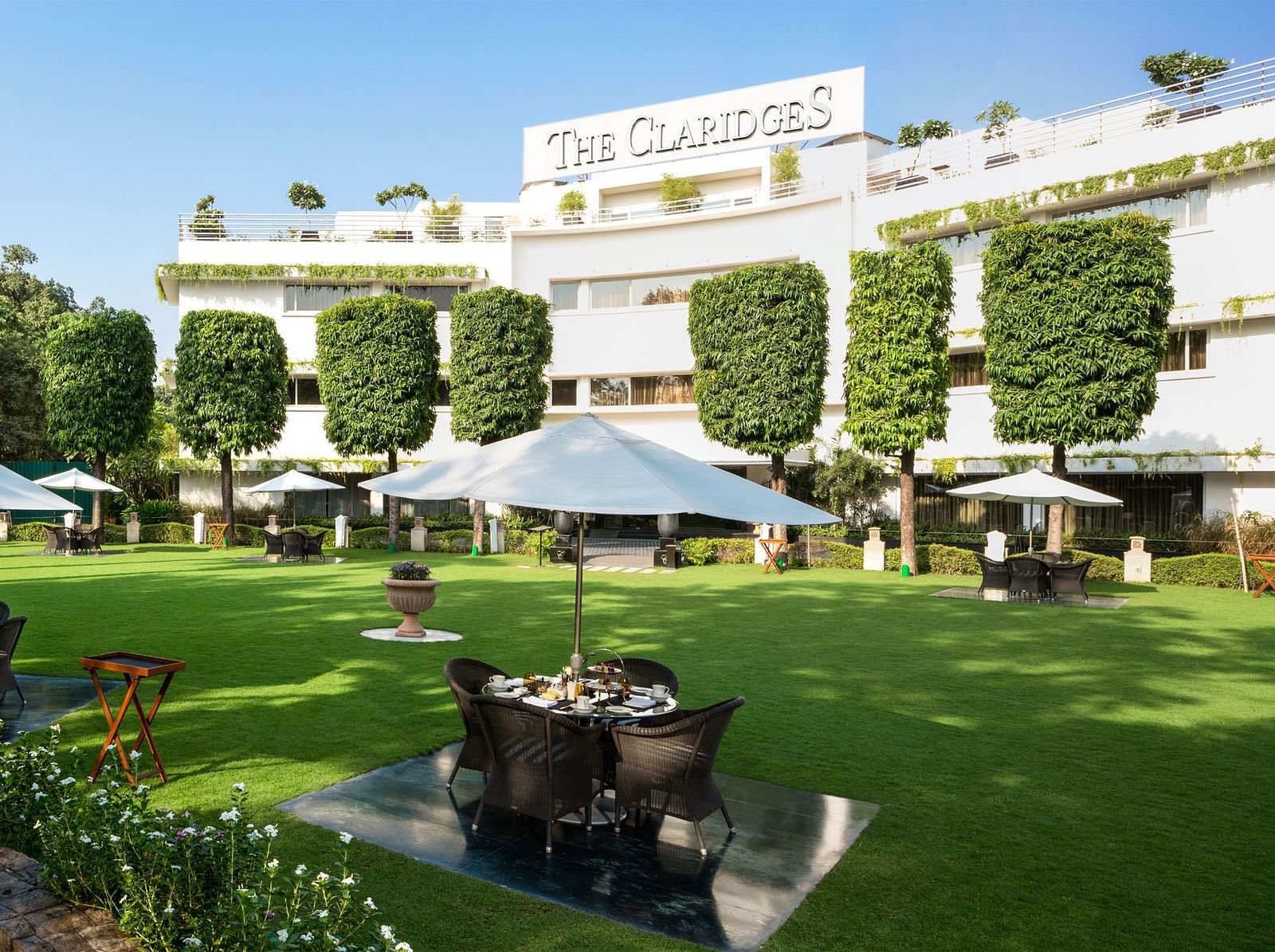 Facade of The Claridges New Delhi, featuring a green garden in the front and surrounded by towering trees, offering a serene and elegant ambience