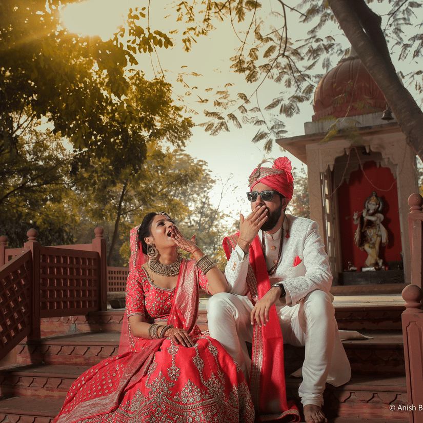 A newlywed couple in wedding attire sits on temple stairs, laughing and playfully covering their mouth at a Jaipur's Top Wedding Resort - Umaid Palace - A Lakeside Heritage Palace Resort