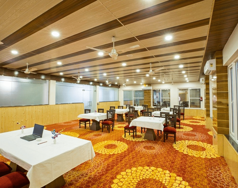 A warmly lit Banquet Hall with a fully carpeted floor along with tables set with white table cloth at DLS Hotels On The Ganges.