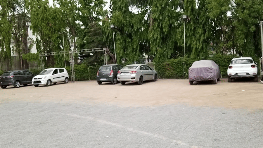 An outdoor open-air gravel parking lot with several cars parked in front of a line of green trees.