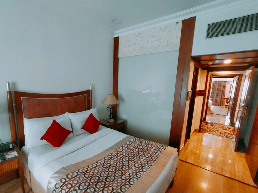 A bedroom at Hotel Hindusthan International, Kolkata, featuring a comfortable bed, bedside tables, and a view leading into a spacious suite area.
