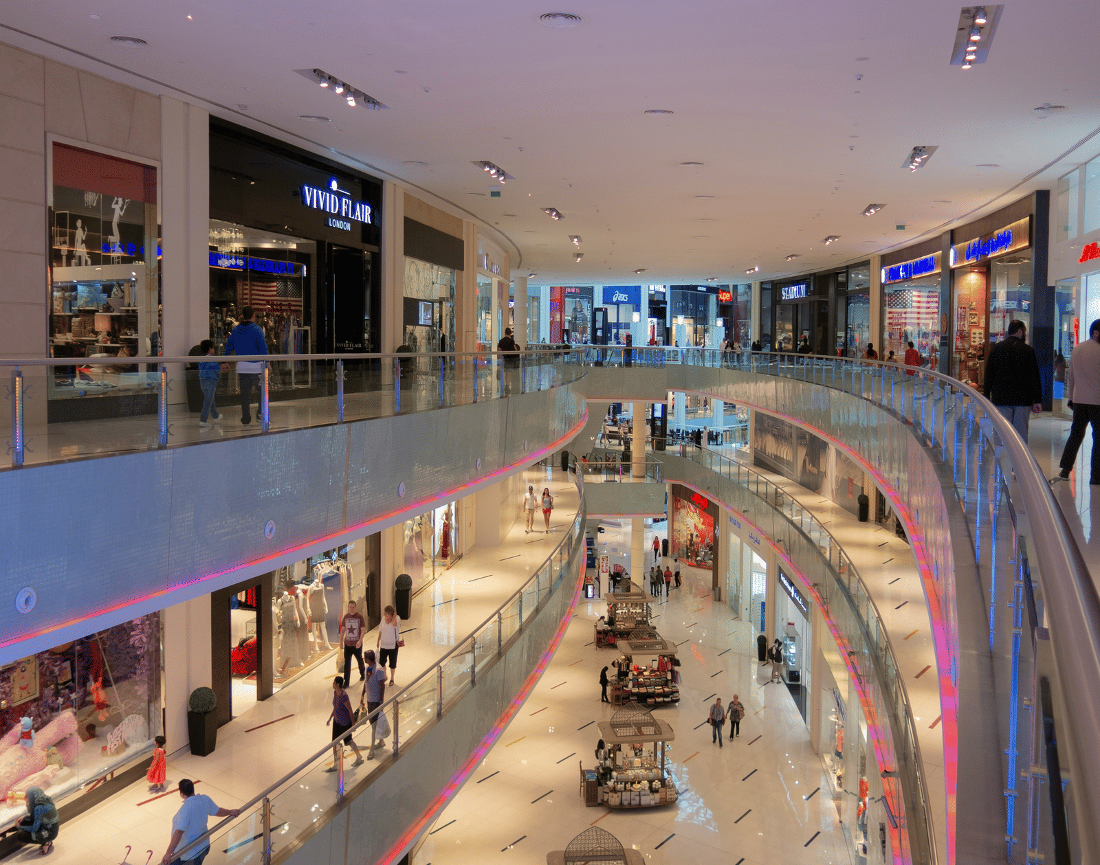 Interior of a modern, multi-level shopping centre with brightly lit shops, glass railings, and escalators visible.