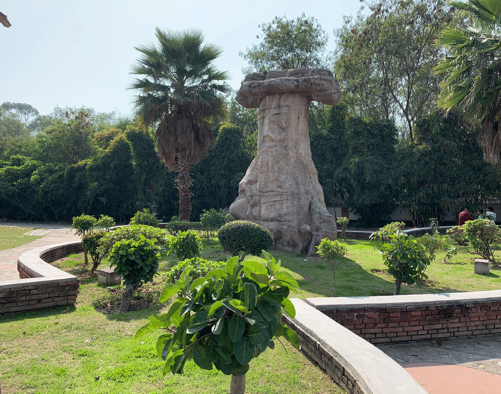 A large, mushroom-shaped stone sculpture stands in a well-maintained public park or garden under a bright, sunny sky.