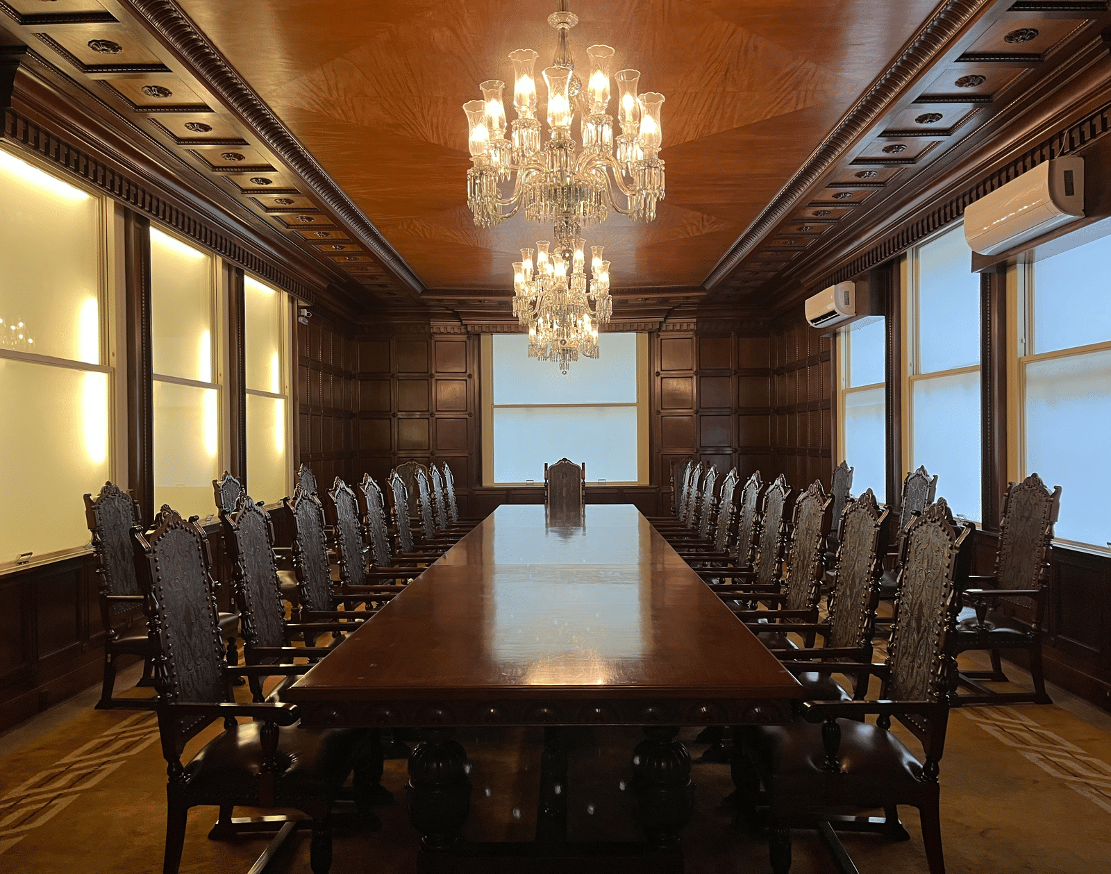 Traditional, luxurious boardroom with dark wooden panelling, a long mahogany table, and crystal chandeliers.