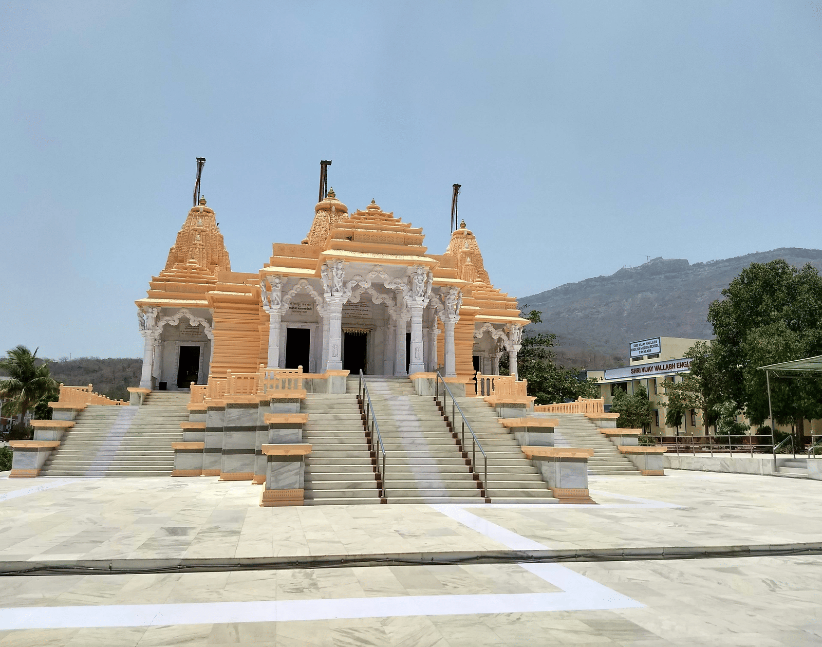 A large, modern temple with peach-coloured sandstone and multiple flights of steps leading to its entrance, set below a hill.
