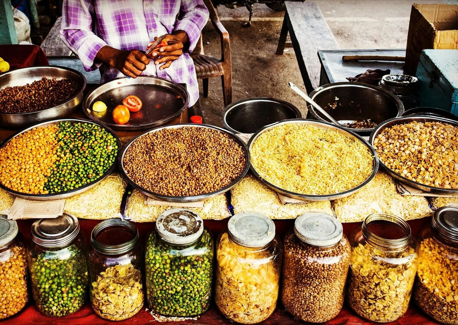 Street food stall with various jars and bowls filled with grains, pulses, and snacks, vendor's hands visible behind.