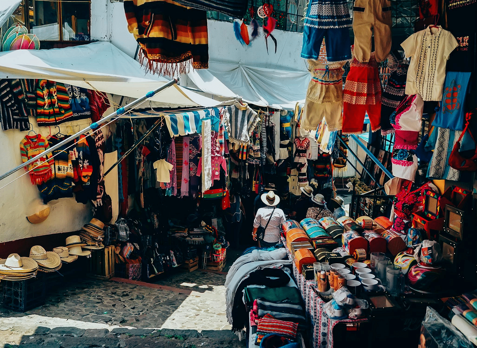 A street market with colorful handmade garments, fabrics, and traditional hats displayed under tents.