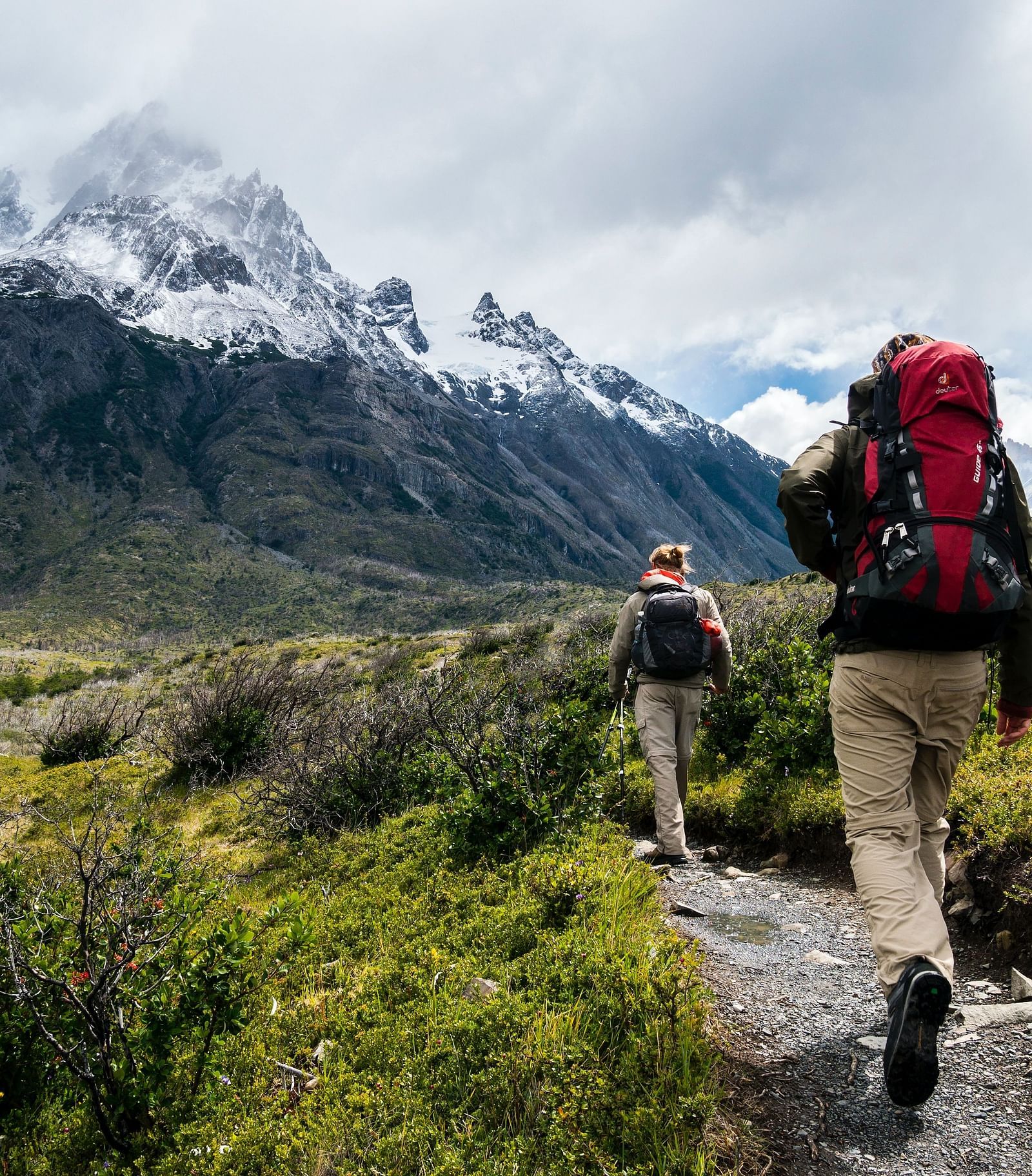 Two people walking on a trekking path with greenery around them and a snow-clad mountain in the background.