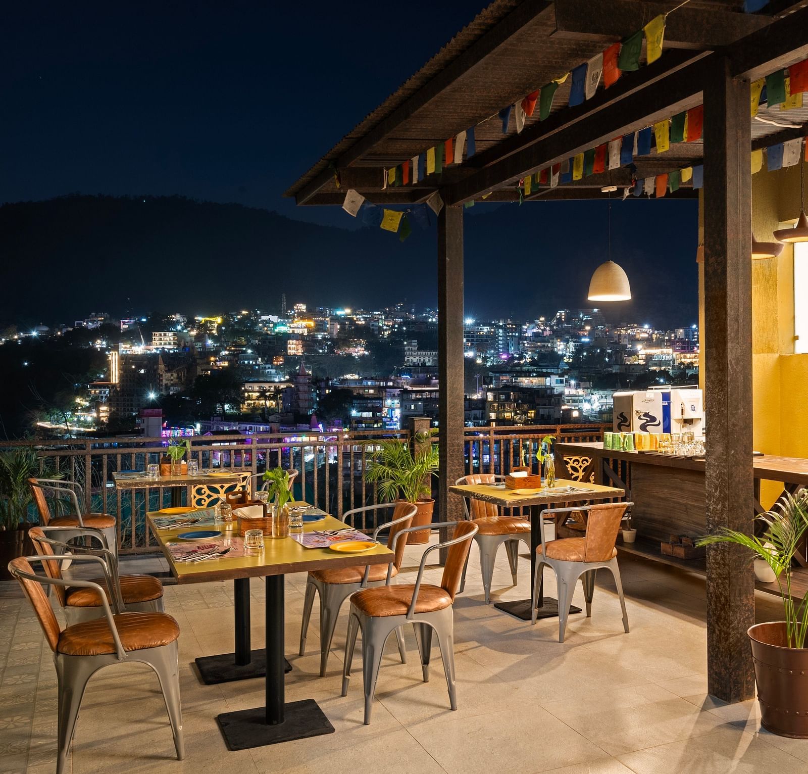 Panoramic view of city skyline from rooftop restaurant, with tables, chairs, and city lights at night at Bedzzz, Rishikesh 