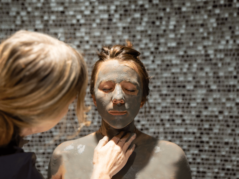 A therapist at YO1 Longevity Health Resorts Catskills applying mud to the face and body of a woman sitting with her eyes closed