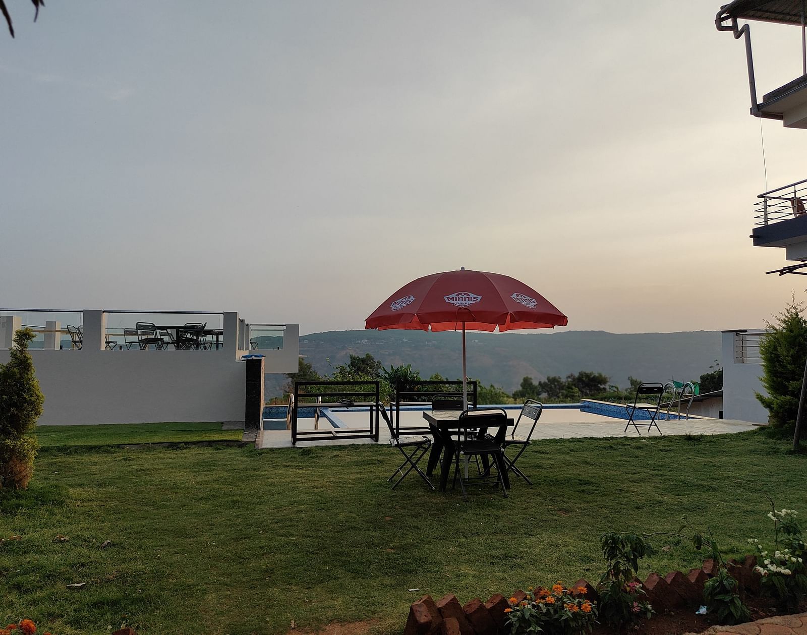 Outdoor area featuring a red umbrella and seating area on a green lawn and a swimming pool, with hills visible in the background.
