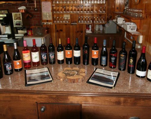 A collection of wine bottles is arranged on a wooden countertop, some with labels visible, along with two framed tasting notes and a wooden coaster, set against a wood-paneled background. - Kit Carson Lodge & Restaurant