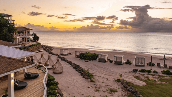 A sunset casts warm hues over a sandy beach leading to the ocean, with a resort visible in the distance.