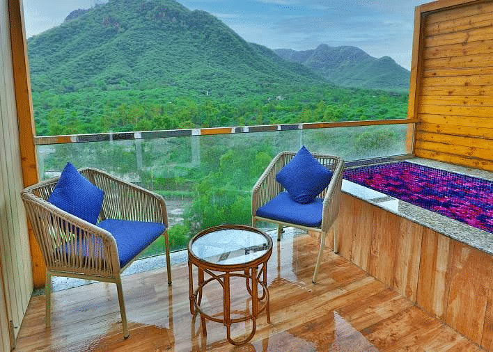 A balcony with two chairs, cushions, and a table overlooks a lush green valley with mountains.- Bamboo Saa Resort & Spa, Udaipur