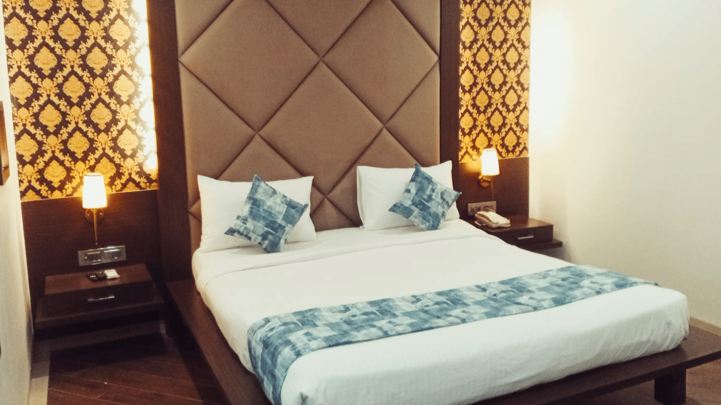 A king size bed with a padded headboard, a patterned wallpaper background, 2 bedside tables, and 2 illuminated wall lamps at Hotel Mumbai House, Thane.