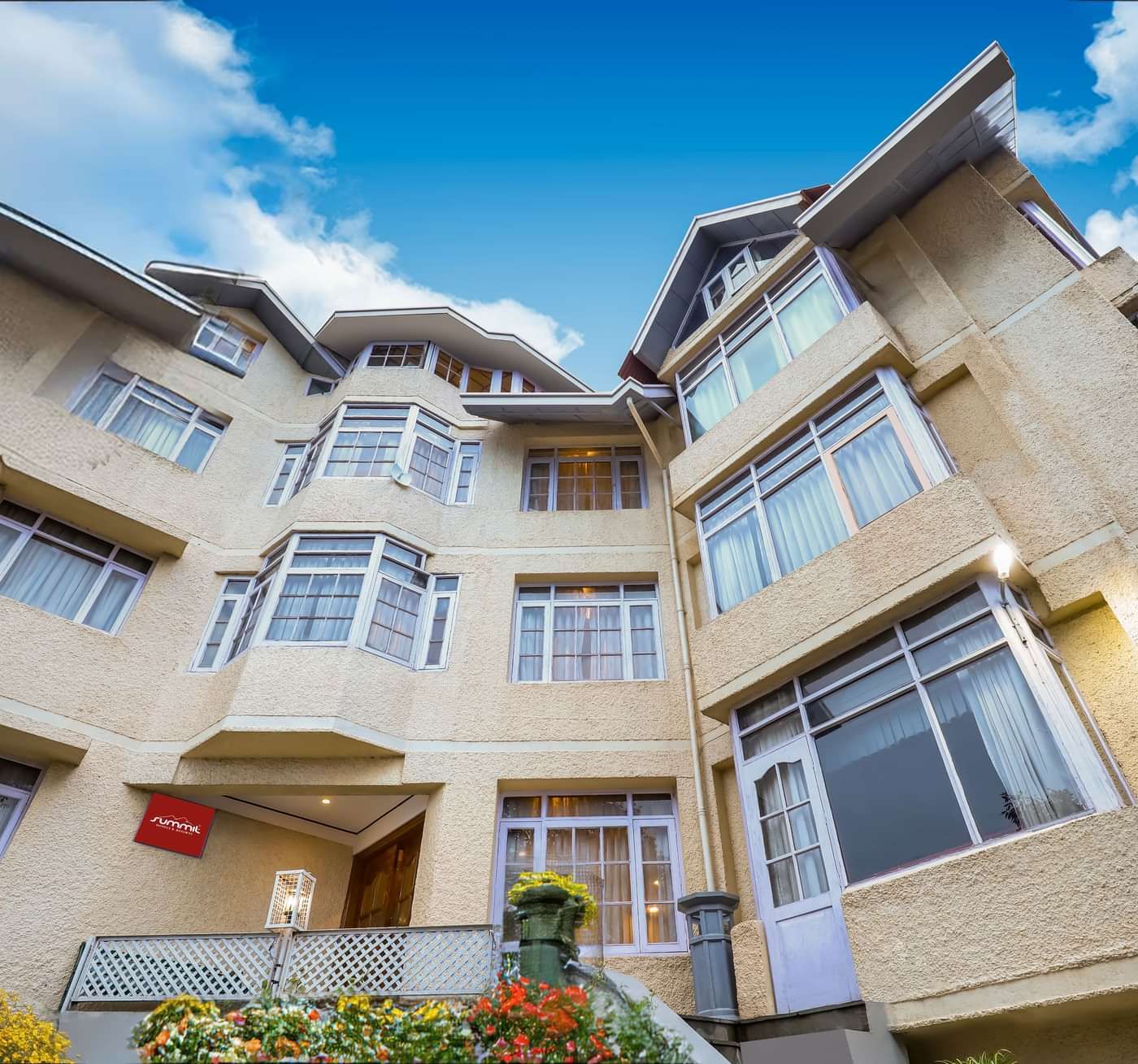 Facade view of Summit Le Royale Hotel, Shimla featuring multiple storey building. Each floor has large windows and the entrance is decorated with plants and flowers. There is also a backdrop of blue and bright sky.