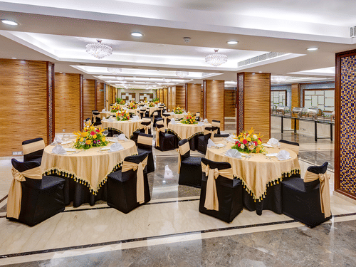 An overview of Regency Hall 1 at Kenilworth Hotel, Kolkata, having round table seating with a black and gold theme, and bouquet, cutlery, and napkins kept on the table.