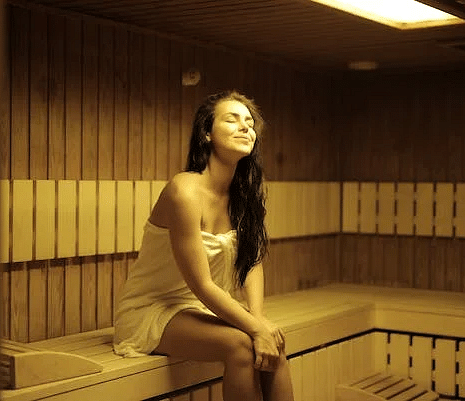 Woman relaxing in a wooden sauna room at YO1, wrapped in a towel, enjoying the warmth and calming ambiance under soft lighting.