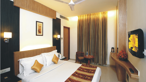 A Deluxe Room at Hotel Southern Regency, Karol Bagh, Delhi, with a bed, warm lighting, full length curtains, a wall-mounted TV, work desk, and seating area.