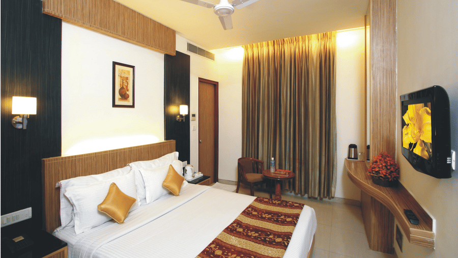 A Deluxe Room at Hotel Southern Regency, Karol Bagh, Delhi, with a bed, warm lighting, full length curtains, a wall-mounted TV, work desk, and seating area.
