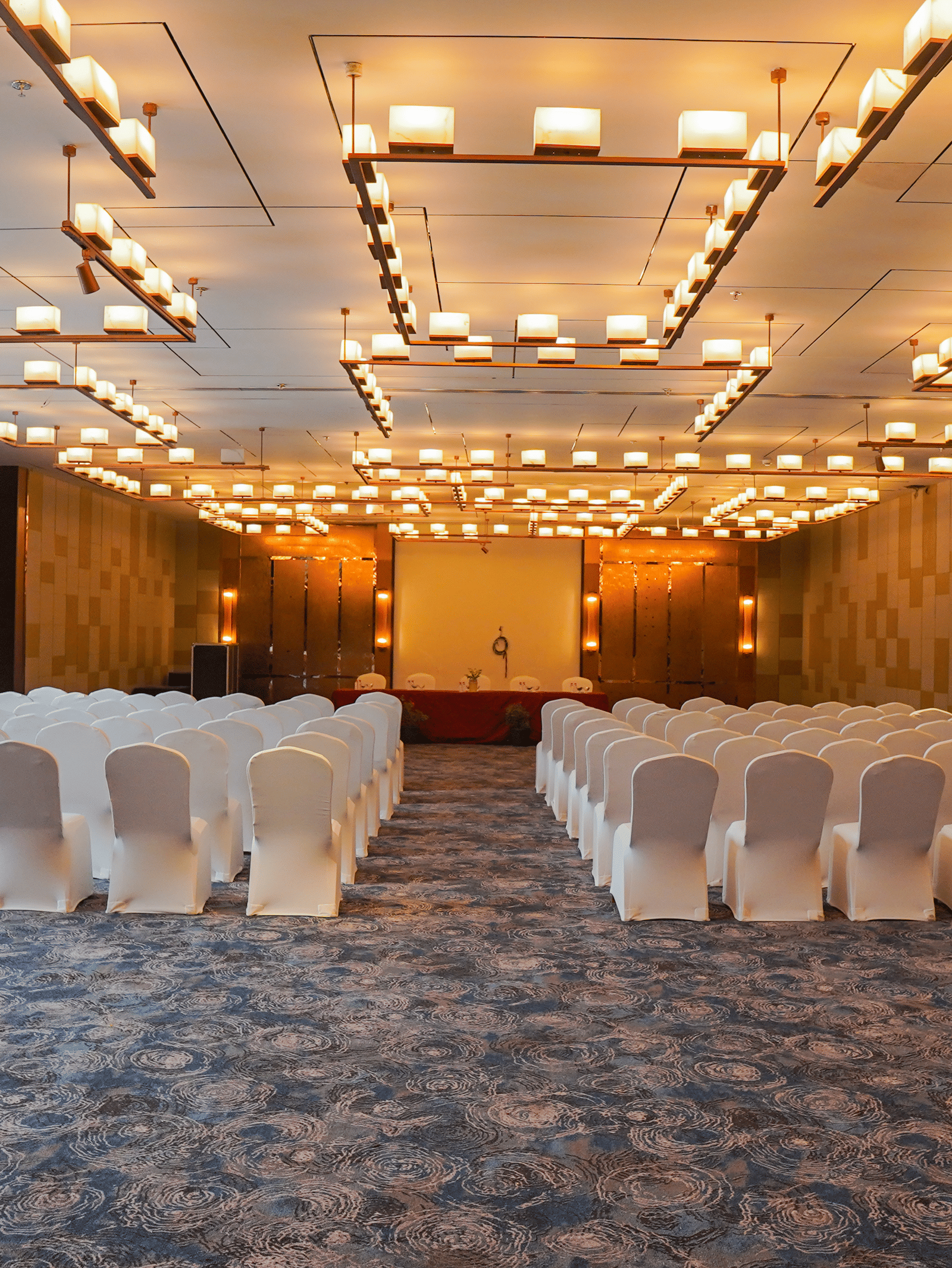 A wide shot of a conference room or ballroom set up with rows of white chairs facing a stage, featuring warm, modern lighting fixtures.