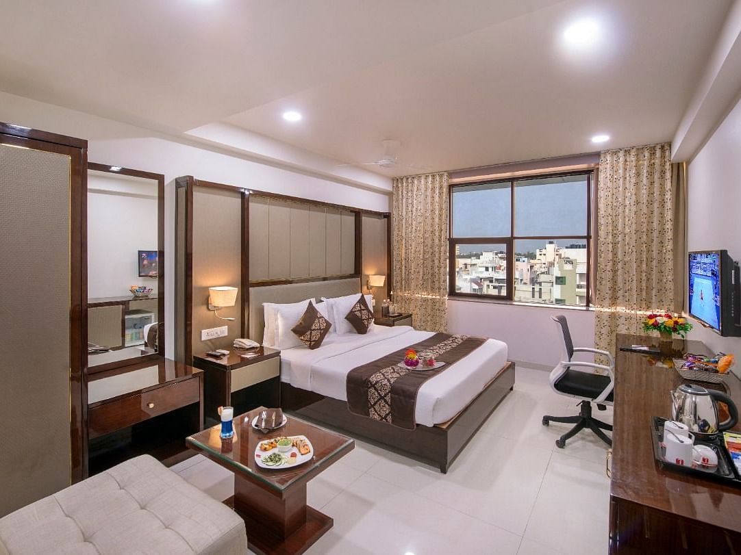 A practical hotel room contains two single beds, a dedicated study desk, a television for guests, and a window showing a cityscape.
