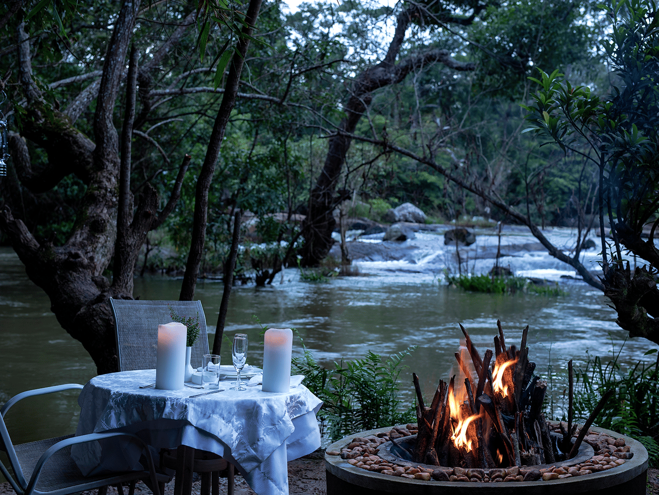 Outdoor candlelit dinner setup by a river in the evening- Amanvana Resort And Spa, Dining in Coorg