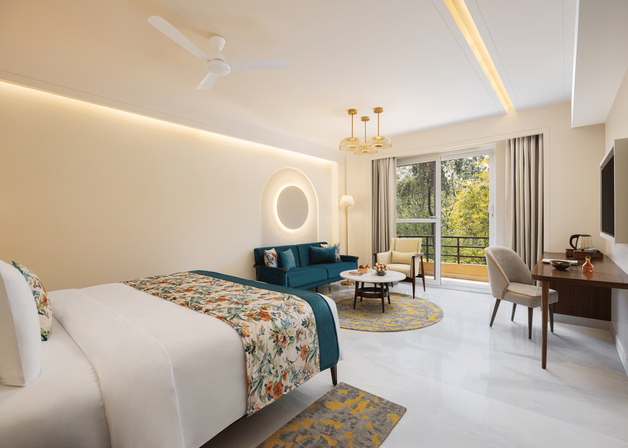 A bright and airy hotel room featuring a queen bed with floral accents, a teal velvet sofa, a round coffee table on a patterned rug, a work desk with a chair and a television, a balcony with outdoor seating and a view of greenery, modern lighting, and a c