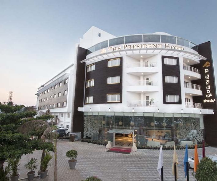 A facade image  with a huge parking area where cars are also parked - The President Hotel,Hubli