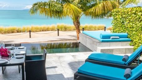 Beachfront patio with turquoise sun loungers, palm trees, and a dining table set against the backdrop of golden sand and calm Caribbean waters.