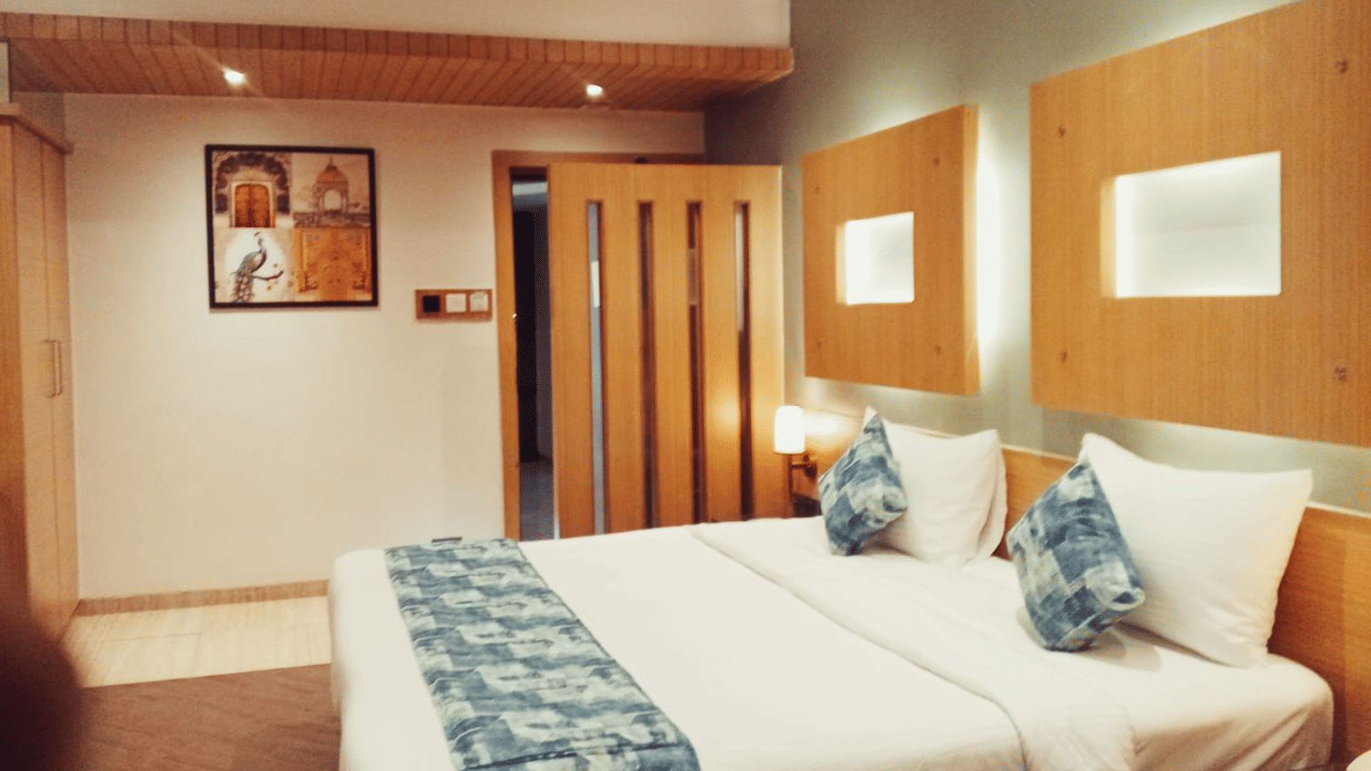 A bedroom space arranged with double beds, a wall-mounted wooden frame, an overhead ceiling fan, and a light-toned wooden wardrobe unit at Hotel Mumbai House, Thane.