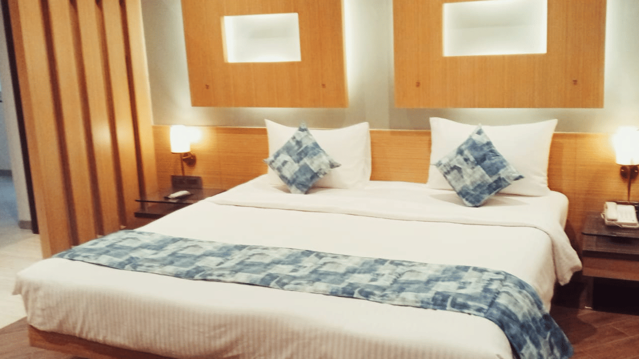 A king size bed with a white duvet, a patterned bed runner, 2 wooden nightstands with lamps, and 2 rectangular wall design panels at Hotel Mumbai House, Thane.
