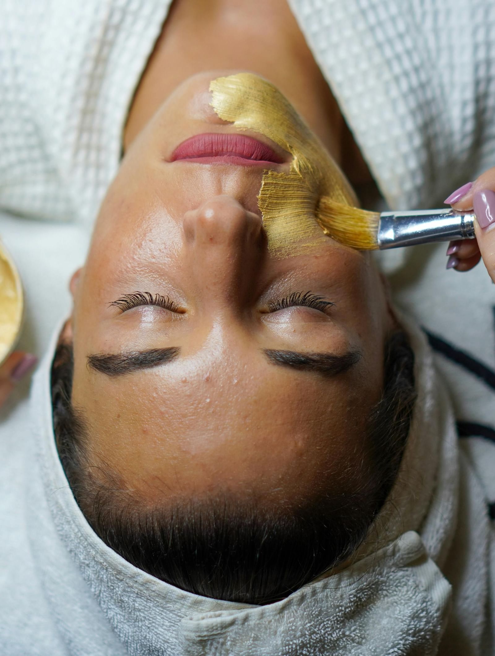 A person receives a facial mask application with a brush, while holding a small bowl of the mask mixture.