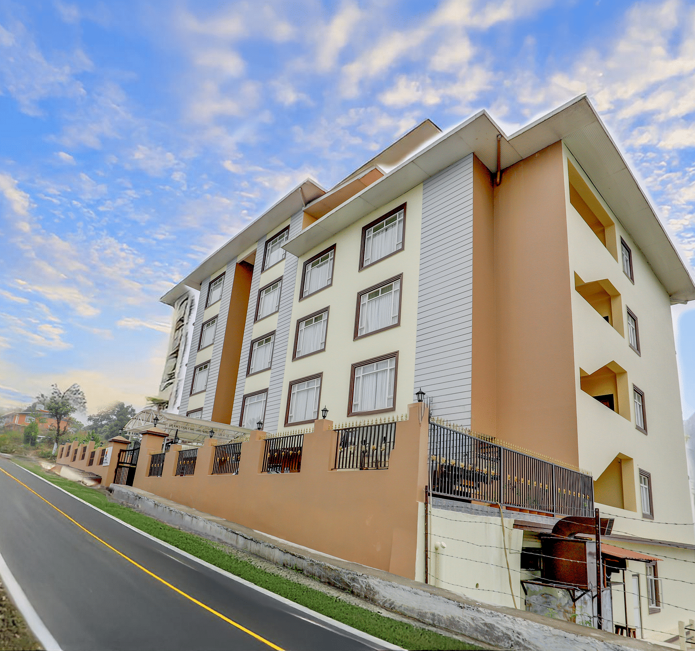 A modern Banari Regency by Summit, Namchi hotel exterior walls stands beside a sloping road, surrounded by greenery and mountains under a blue sky with scattered clouds